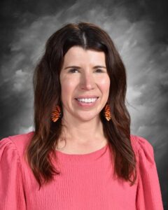 Smiling woman with long brown hair, wearing orange butterfly earrings and a pink top, against a gray background.