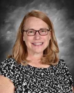 Smiling woman with shoulder-length blonde hair, wearing glasses and a black floral blouse, against a gray background.
