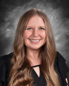 A woman with long, wavy hair smiles warmly, wearing a black top against a gray backdrop.