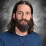 Un hombre con el pelo largo y oscuro y barba, vestido con una camisa azul y un collar, aparece de pie sobre un fondo gris.