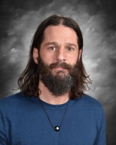 A man with long dark hair and a beard, wearing a blue shirt and necklace, stands against a gray background.