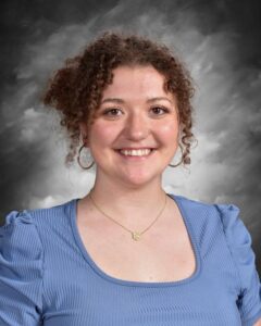 Smiling young woman with curly hair, wearing a blue top and a necklace, against a gray background.