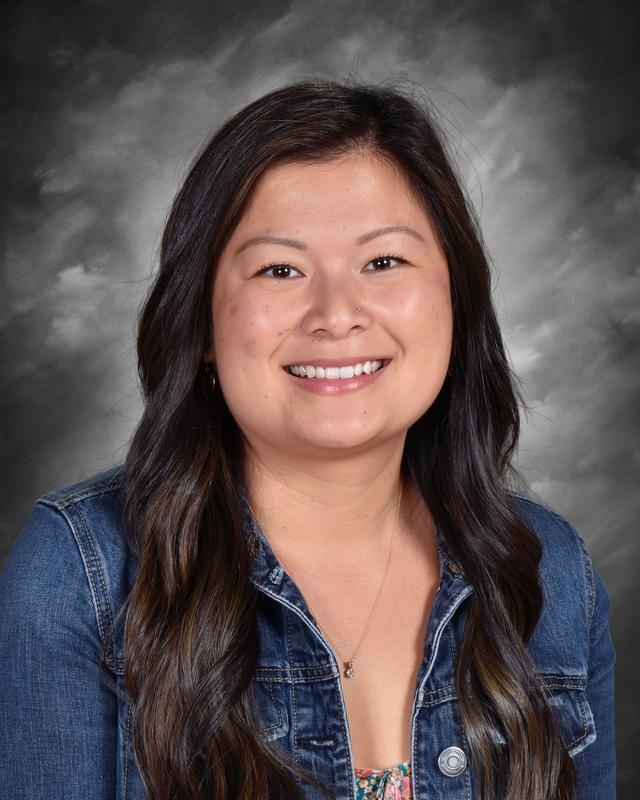 A smiling woman with long dark hair wearing a denim jacket, set against a gray backdrop.