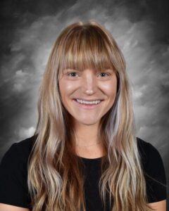 Smiling woman with long, wavy hair, wearing a black shirt, against a gray background.