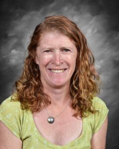 Smiling woman with curly red hair wearing a green shirt and silver necklace against a gray background.