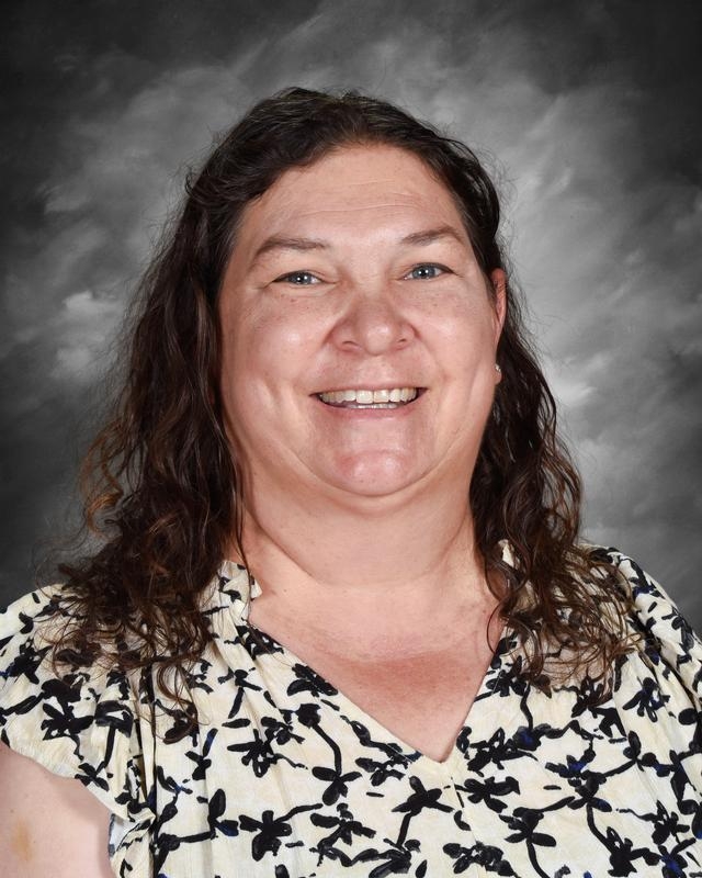 Smiling woman with long, curly hair wearing a floral blouse, set against a gray background.