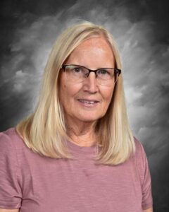 A woman with long blonde hair and glasses smiles, wearing a pink shirt against a soft gray background.