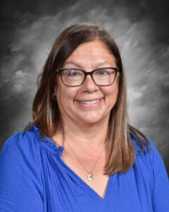 Smiling woman with long brown hair wearing glasses and a blue blouse, set against a gray background.