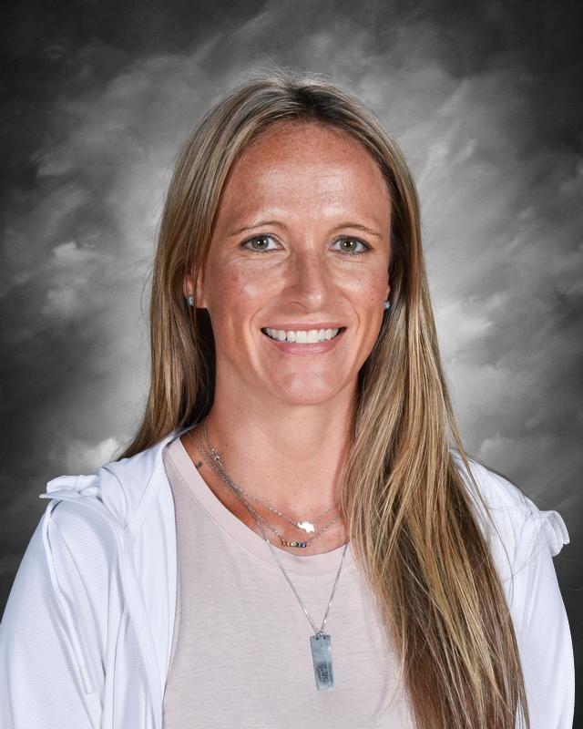 A woman with long blonde hair smiles, wearing a light top and layered necklaces, set against a gray background.