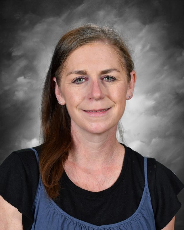 A woman with long brown hair smiles, wearing a black shirt and blue top, against a gray background.