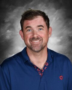 Smiling man with short brown hair, wearing a blue polo shirt, against a gray background.