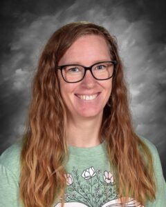 Smiling woman with long, wavy hair wearing glasses and a green t-shirt with a floral design. Gray background.