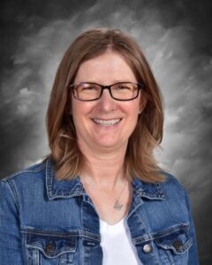 Smiling woman with glasses and shoulder-length hair, wearing a denim jacket over a white shirt, against a gray background.