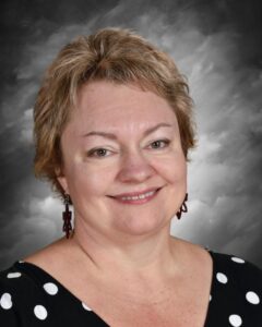 Smiling woman with short, light brown hair, wearing a black polka dot dress and red earrings, against a gray background.