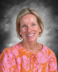 Smiling woman with short blonde hair, wearing a bright pink and orange floral top, against a gray background.