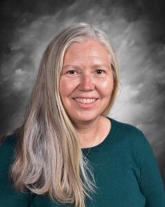 A woman with long, straight, gray hair smiles, wearing a dark green sweater against a soft gray background.
