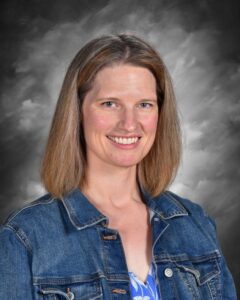 Smiling woman with shoulder-length brown hair, wearing a denim jacket over a blue patterned top, against a gray backdrop.