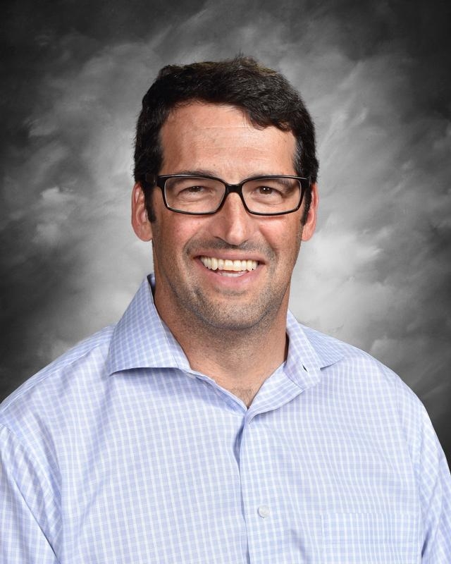 Smiling man with glasses and short dark hair, wearing a light blue checkered shirt, against a gray background.