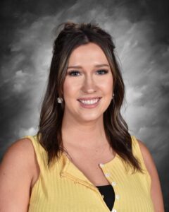 A smiling woman with long brown hair and a yellow top poses against a gray background.