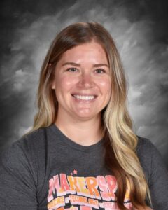 Smiling woman with long, wavy hair wearing a gray t-shirt with colorful text, set against a cloudy background.