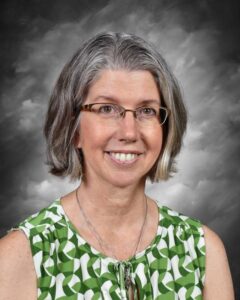 A woman with gray hair and glasses smiles, wearing a green patterned blouse against a gray background.