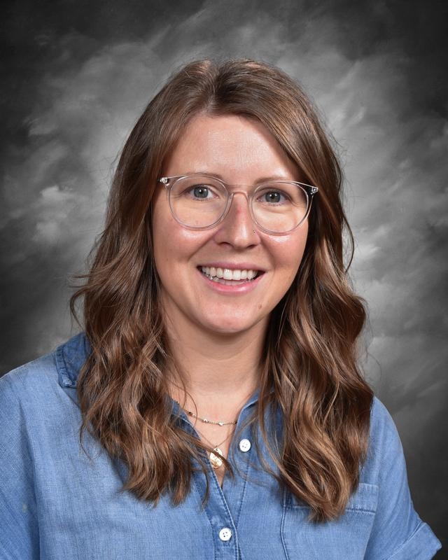 Smiling woman with wavy brown hair, wearing glasses and a blue shirt, against a gray background.