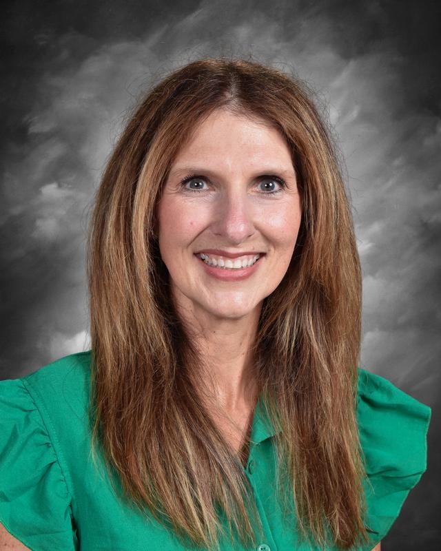 A smiling woman with long brown hair, wearing a green blouse, against a gray background.