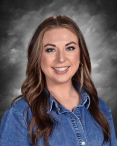 Smiling woman with long, wavy brown hair wearing a denim shirt, set against a gray background.
