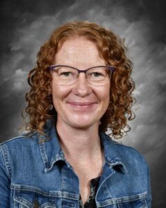 A woman with curly red hair, wearing glasses and a denim jacket, smiles against a gray background.
