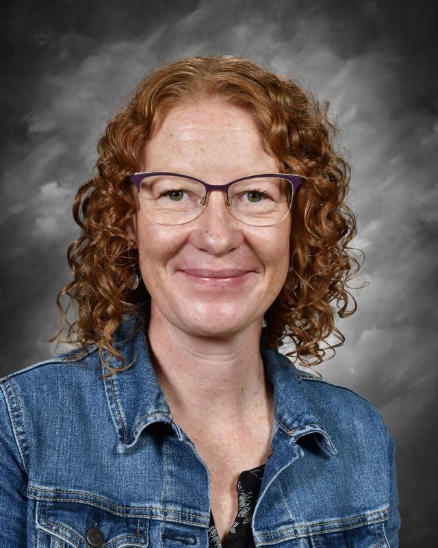 A woman with curly red hair, wearing glasses and a denim jacket, smiles against a gray background.