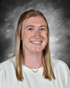 Smiling young woman with long blonde hair, wearing a white sweater and colorful earrings, against a gray background.