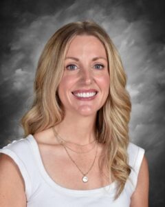 Mujer sonriente con el pelo largo y rubio ondulado, vestida con una camiseta sin mangas blanca y varios collares, sobre un fondo gris.