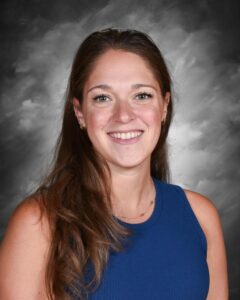 Smiling woman with long brown hair wearing a blue sleeveless top, set against a gray background.