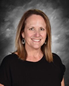 A woman with shoulder-length brown hair smiles, wearing a black shirt and dangling earrings, against a gray background.