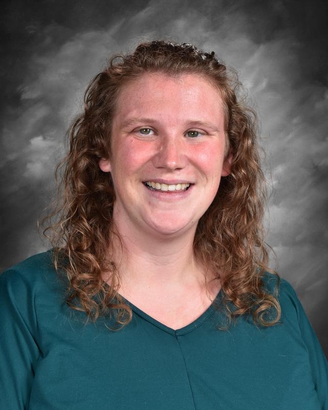 Smiling woman with curly hair, wearing a teal top, against a gray background.
