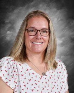 Smiling woman with shoulder-length blonde hair, wearing glasses and a white shirt with cherry patterns, against a gray backdrop.