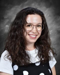 Smiling young woman with curly hair, wearing glasses and a polka dot dress over a white shirt, against a gray background.