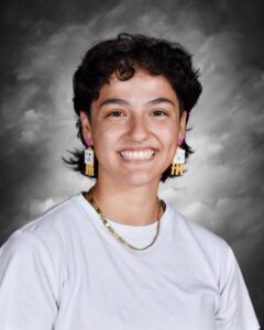A smiling person with short, curly hair wearing colorful earrings and a white shirt against a gray background.