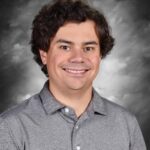 Smiling man with curly hair wearing a gray polo shirt, set against a gray background.
