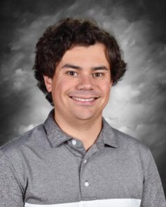 Smiling man with curly hair wearing a gray polo shirt, set against a gray background.