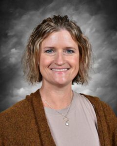 Smiling woman with short, wavy hair, wearing a brown cardigan and necklace, against a cloudy gray background.