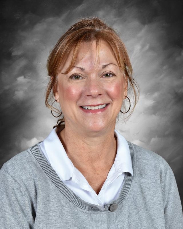 Smiling woman with shoulder-length brown hair, wearing a gray sweater over a white shirt, against a gray background.