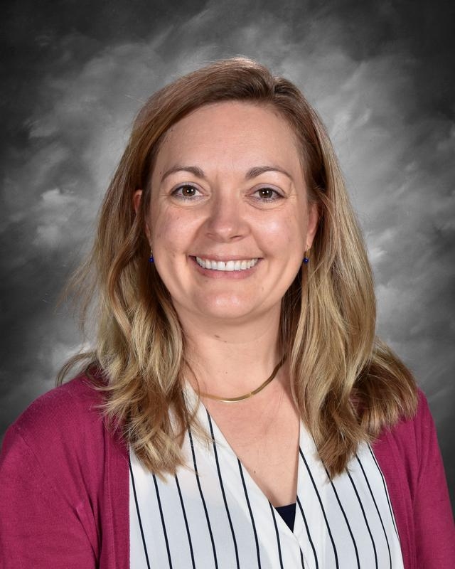 Smiling woman with long, wavy hair, wearing a striped shirt and a maroon cardigan, against a gray background.