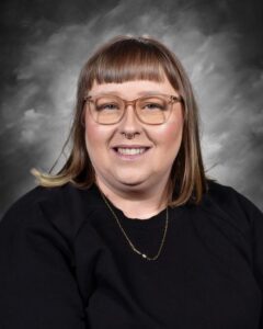 A woman with shoulder-length hair and glasses smiles warmly, wearing a black sweater against a gray background.