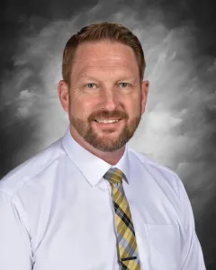 Smiling man with a beard in a white shirt and plaid tie, set against a gray cloudy background.