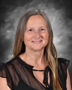 Smiling woman with long hair, wearing a sheer black blouse, in front of a gray, cloudy backdrop.