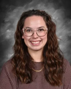 Smiling person with long wavy hair and glasses, wearing a striped sweater and beaded necklace against a gray background.