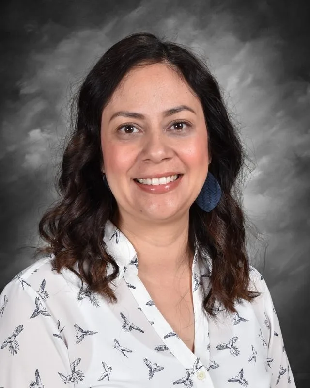 Smiling woman with dark hair, wearing a white shirt with bird prints and blue earrings, against a cloudy gray background.
