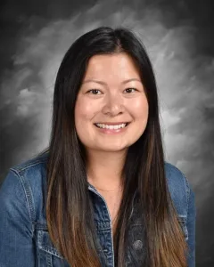 Smiling person with long dark hair in a denim jacket against a gray background.