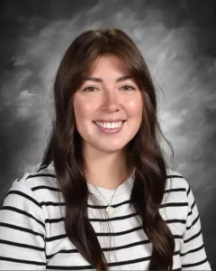 Smiling woman with long brown hair wearing a striped shirt, in front of a cloudy gray background.
